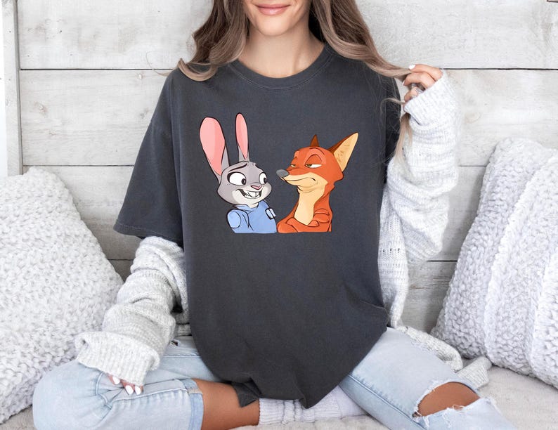 May include: Dark gray t-shirt featuring a cartoon illustration of a rabbit and a fox. The rabbit has gray fur and pink ears, and the fox is orange. The characters are from the animated movie Zootopia.
