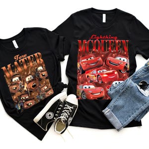 May include: Two black t-shirts with graphic designs, one featuring Tow Mater and the other Lightning McQueen from the Cars movie. The image also includes a pair of blue jeans, black sneakers, and sunglasses.