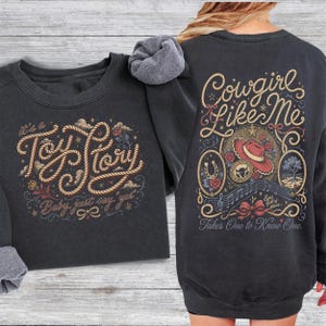 May include: A charcoal gray crewneck sweatshirt featuring a front graphic with rope-style lettering that reads "It's a Toy Story, Baby, just say yes". The back graphic displays "Cowgirl Like Me" with a cowboy hat, horseshoe, and musical notes.