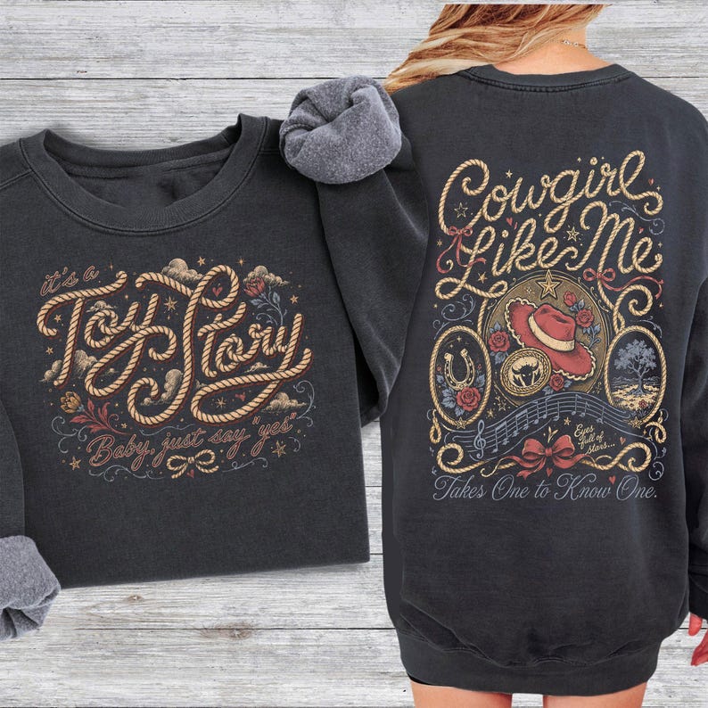 May include: A charcoal gray crewneck sweatshirt featuring a front graphic with rope-style lettering that reads "It's a Toy Story, Baby, just say yes". The back graphic displays "Cowgirl Like Me" with a cowboy hat, horseshoe, and musical notes.