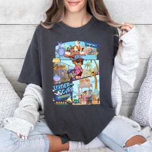 May include: Dark gray t-shirt featuring a colorful graphic from the animated movie "Undercover Mission." The design includes cartoon characters in sunglasses, a beach scene, and the words "Undercover Mission" and "Big Pop."