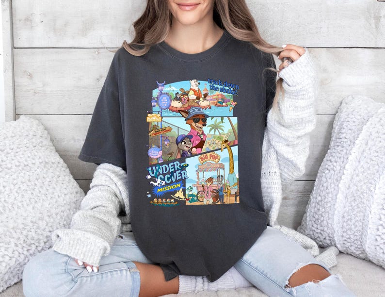 May include: Dark gray t-shirt featuring a colorful graphic from the animated movie "Undercover Mission." The design includes cartoon characters in sunglasses, a beach scene, and the words "Undercover Mission" and "Big Pop."