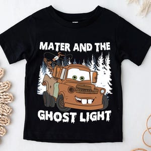 May include: Black short-sleeved t-shirt with a cartoon image of Mater from the film Cars. The image shows Mater in brown and tan with a forest backdrop. The text "Mater and the Ghost Light" is printed above and below the image in white lettering.