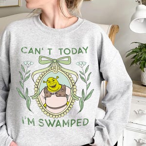 Puede incluir: Sudadera gris con un personaje de dibujos animados de ogro verde en un marco con el texto "Can't Today I'm Swamped".