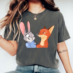 May include: Dark grey t-shirt featuring a cartoon illustration of a smiling rabbit and a sly fox. The rabbit has pink ears and a blue uniform, while the fox is orange and red. The shirt is a comfortable, casual style.