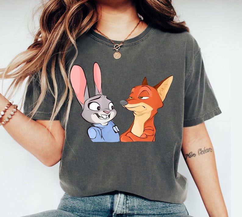 May include: Dark grey t-shirt featuring a cartoon illustration of a smiling rabbit and a sly fox. The rabbit has pink ears and a blue uniform, while the fox is orange and red. The shirt is a comfortable, casual style.
