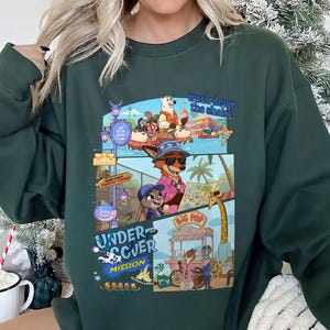 May include: Dark green sweatshirt featuring a colorful graphic from the movie Zootopia. The graphic includes the text "track down the shake!" and "Undercover Mission" with various cartoon characters and scenes. The sweatshirt is a crew neck style.