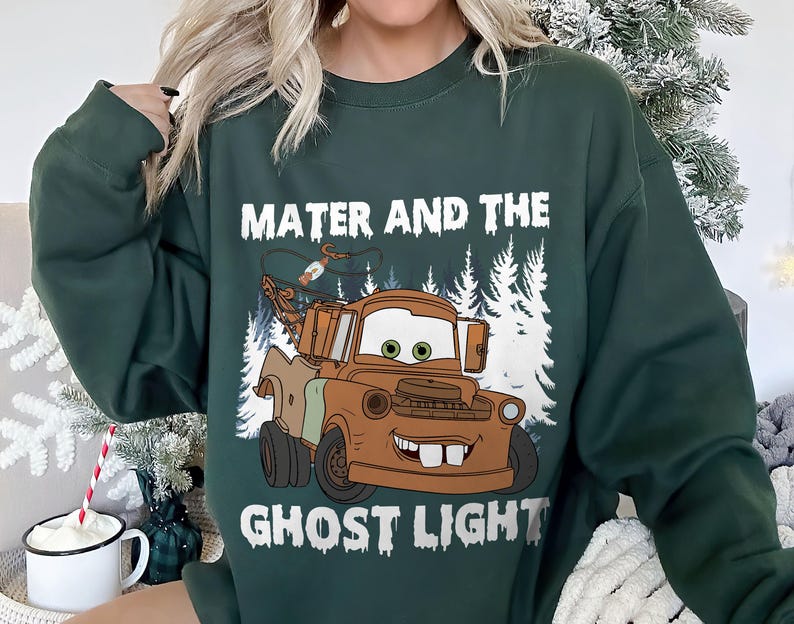 May include: Dark green sweatshirt with a cartoon image of Mater from the film Cars, and the text "Mater and the Ghost Light" in white. The image shows Mater as a brown tow truck with a smiling face, set against a backdrop of white trees.
