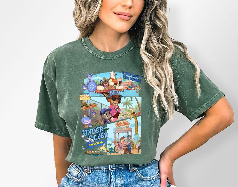 May include: Green t-shirt featuring a colorful graphic of cartoon characters in a beach scene. The graphic includes the text "Undercover Mission" and "Big Pop". The shirt is made of a soft material.