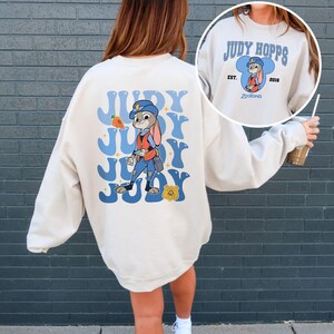 May include: Cream-colored sweatshirt featuring a cartoon image of Judy Hopps from Zootopia. The back design includes the name "JUDY" in blue bubble letters with a carrot and badge graphic. The front has a smaller design with Judy Hopps' name and the year 2016.