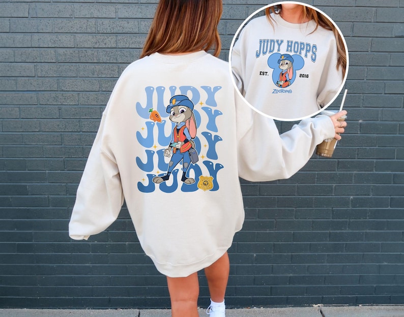 May include: Cream-colored sweatshirt featuring a cartoon image of Judy Hopps from Zootopia. The back design includes the name "JUDY" in blue bubble letters with a carrot and badge graphic. The front has a smaller design with Judy Hopps' name and the year 2016.