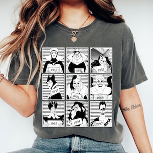 May include: Dark gray t-shirt featuring a grid of nine black and white illustrations of Disney villains, each with a year. The shirt has a crew neck and short sleeves. The illustrations include the Evil Queen, Cruella de Vil, and Ursula.