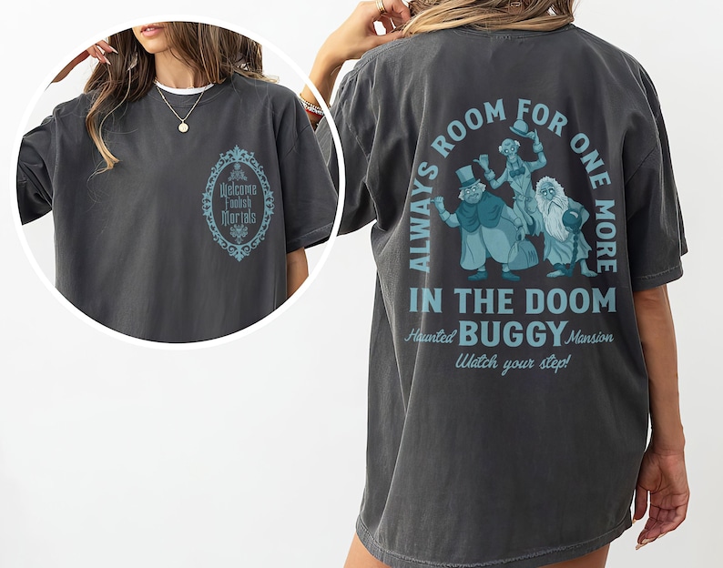 May include: Dark gray t-shirt with a teal graphic on the back featuring three cartoon figures and the text "ALWAYS ROOM FOR ONE MORE IN THE DOOM BUGGY Mansion". The front of the shirt has a teal oval design with the words "Welcome Foolish Mortals".