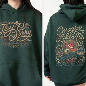 May include: A dark green hooded sweatshirt featuring a graphic on the front that reads "It's a Toy Story, Baby, just say yes" in rope-style lettering. The back displays "Cowgirl Like Me" and "Takes One to Know One" with a cowboy hat and floral art.
