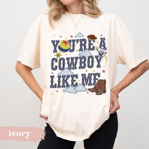 May include: An ivory short-sleeve t-shirt featuring the text "YOU'RE A COWBOY LIKE ME" in dark blue letters. The graphic includes a yellow, red, and blue ball, a brown cowboy hat, a rocket ship, brown cowboy boots, and small colorful stars.