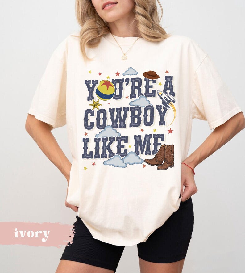 May include: An ivory short-sleeve t-shirt featuring the text "YOU'RE A COWBOY LIKE ME" in dark blue letters. The graphic includes a yellow, red, and blue ball, a brown cowboy hat, a rocket ship, brown cowboy boots, and small colorful stars.