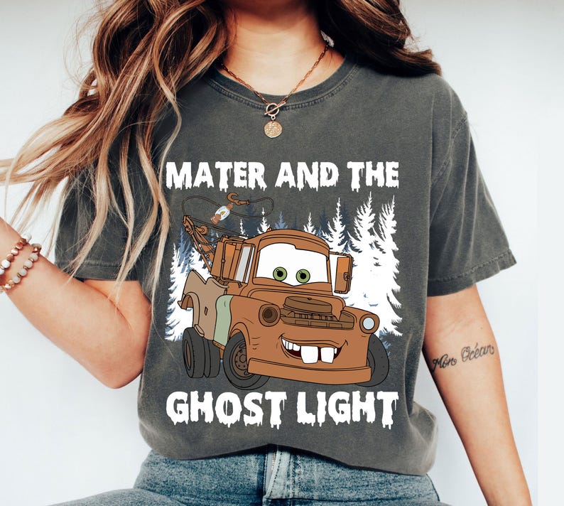 May include: Dark grey t-shirt with a cartoon image of Mater from the film Cars, and the text "Mater and the Ghost Light" in white. The graphic shows a brown tow truck with green eyes and a smiling mouth, set against a backdrop of white trees.