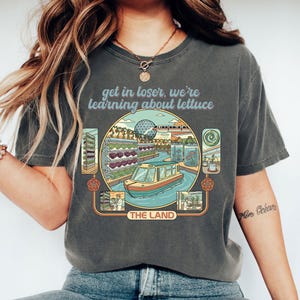 Könnte beinhalten: Dunkelgraues T-Shirt mit einer Grafik einer Bootsfahrt durch eine Hydrokulturfarm, mit dem Text "get in loser, we're learning about lettuce" über der Grafik und "THE LAND" darunter.
