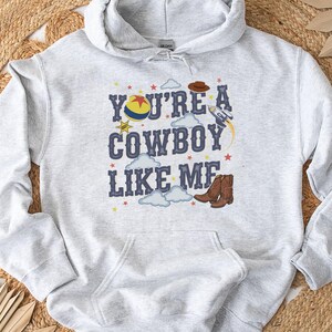 May include: A light gray hooded sweatshirt featuring a graphic print with the text "YOU'RE A COWBOY LIKE ME" in dark blue letters. The design includes a colorful ball, a brown cowboy hat, a rocket ship, clouds, stars, and brown cowboy boots.