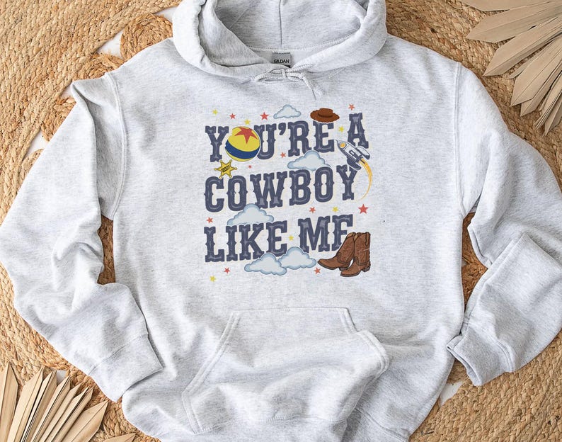 May include: A light gray hooded sweatshirt featuring a graphic print with the text "YOU'RE A COWBOY LIKE ME" in dark blue letters. The design includes a colorful ball, a brown cowboy hat, a rocket ship, clouds, stars, and brown cowboy boots.