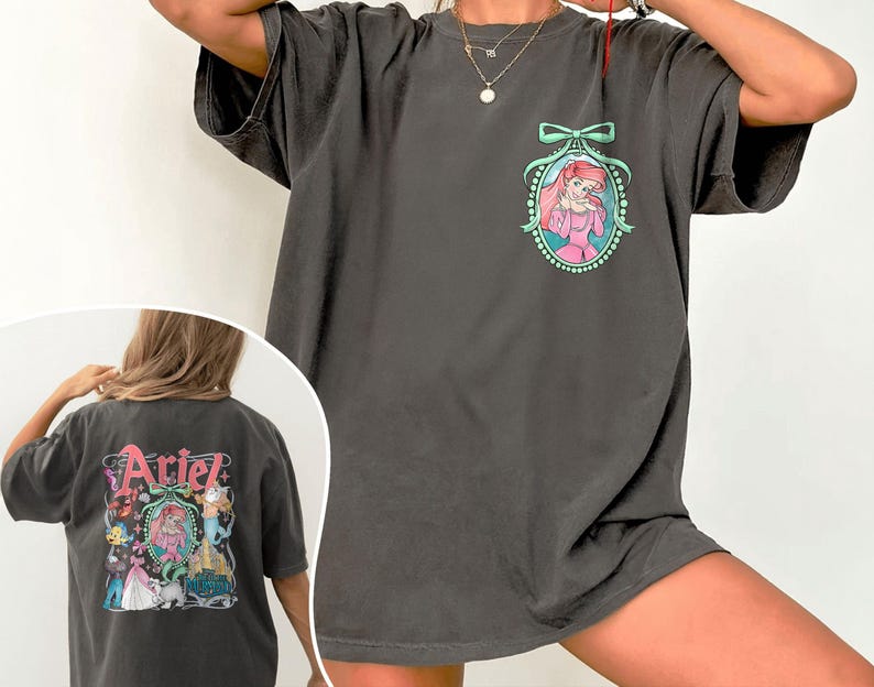 Pu&ograve; includere: Una maglietta grigia oversize con una stampa grafica di Ariel de La Sirenetta. La grafica presenta Ariel con un vestito rosa, un fiocco verde e una collana di perle. Il testo "Ariel" &egrave; stampato sul retro della maglietta.