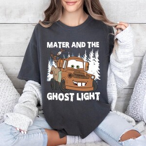 May include: Dark grey t-shirt with a cartoon image of Mater from the film Cars, and the text "Mater and the Ghost Light" in white. The graphic shows a brown truck with a smiling face, set against a backdrop of white trees.