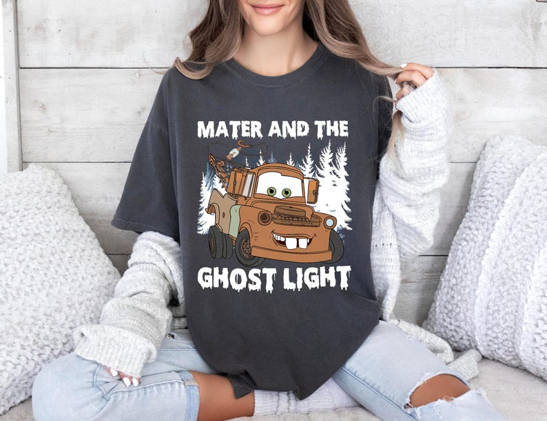 May include: Dark grey t-shirt with a cartoon image of Mater from the film Cars, and the text "Mater and the Ghost Light" in white. The graphic shows a brown truck with a smiling face, set against a backdrop of white trees.