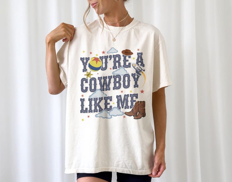 May include: An off-white short-sleeve t-shirt featuring the blue text "YOU'RE A COWBOY LIKE ME" surrounded by graphic illustrations of a yellow and blue ball, a brown cowboy hat, a rocket ship, a sheriff star, brown cowboy boots, and clouds.