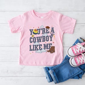 May include: A light pink short-sleeve t-shirt featuring the text "YOU'RE A COWBOY LIKE ME" in dark blue letters. The graphic includes a colorful ball, a brown cowboy hat, a rocket ship, brown cowboy boots, small stars, and white clouds.