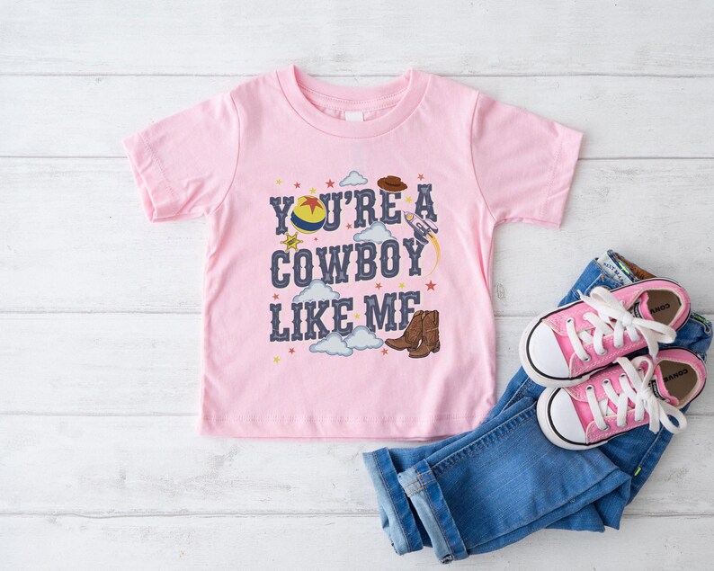 May include: A light pink short-sleeve t-shirt featuring the text "YOU'RE A COWBOY LIKE ME" in dark blue letters. The graphic includes a colorful ball, a brown cowboy hat, a rocket ship, brown cowboy boots, small stars, and white clouds.