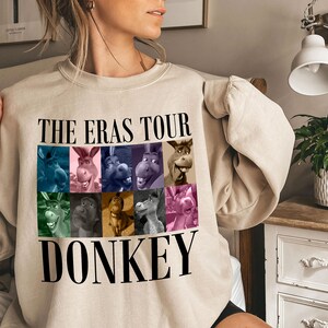 Pode incluir: Uma camisa de moletom bege com o texto "THE ERAS TOUR DONKEY" estampado na frente. O texto &eacute; preto e h&aacute; nove imagens coloridas de um burro em um padr&atilde;o de grade acima do texto.
