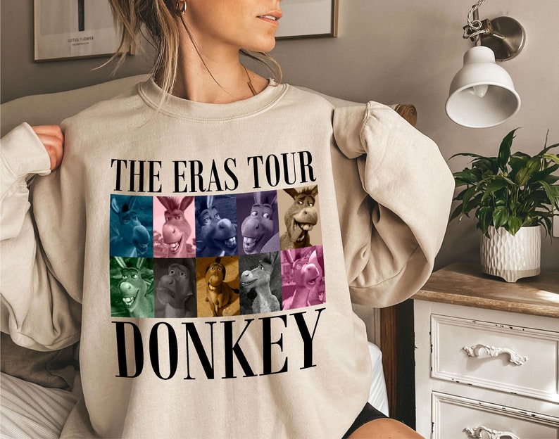 Pode incluir: Uma camisa de moletom bege com o texto "THE ERAS TOUR DONKEY" estampado na frente. O texto &eacute; preto e h&aacute; nove imagens coloridas de um burro em um padr&atilde;o de grade acima do texto.