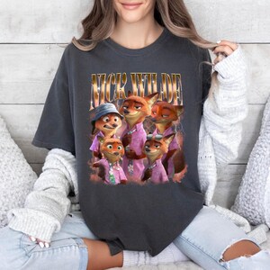 May include: Dark gray t-shirt featuring a graphic of the characters from the animated movie Zootopia. The text "Nick Wilde" is printed above the image of the fox characters. The characters are wearing pink shirts.