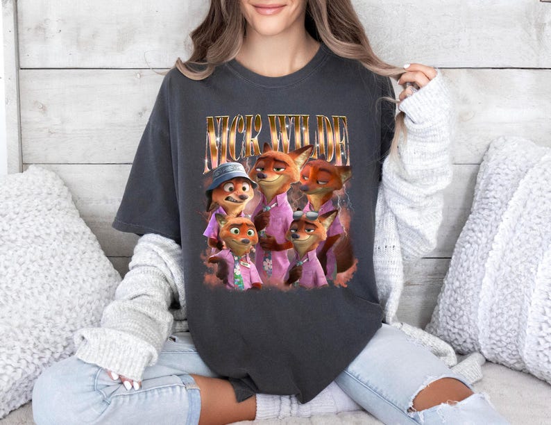 May include: Dark gray t-shirt featuring a graphic of the characters from the animated movie Zootopia. The text "Nick Wilde" is printed above the image of the fox characters. The characters are wearing pink shirts.