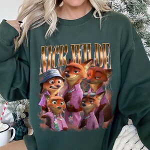 May include: Dark green sweatshirt featuring a graphic of Nick Wilde from Zootopia. The image shows Nick Wilde and other fox characters in pink shirts. The text "NICK WILDE" is printed above the characters in a gold, glittery font.