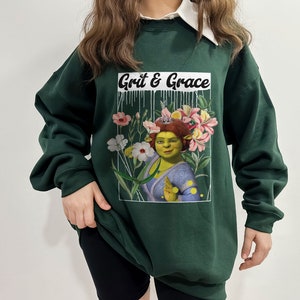 May include: A dark green sweatshirt with a white collar and a graphic print of a green ogre with flowers and the text "Grit & Grace".