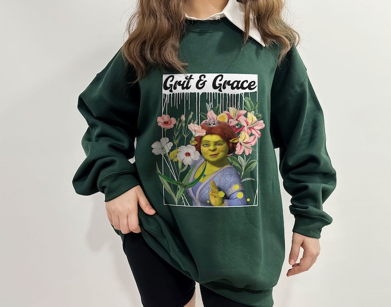 May include: A dark green sweatshirt with a white collar and a graphic print of a green ogre with flowers and the text "Grit & Grace".