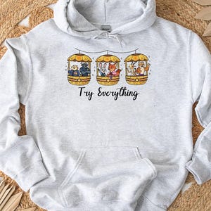 Pode incluir: Moletom cinza claro com bolso canguru e capuz com cord&atilde;o. A frente apresenta um gr&aacute;fico de tr&ecirc;s g&ocirc;ndolas, cada uma com um animal, e as palavras "Try Everything" em escrita preta.