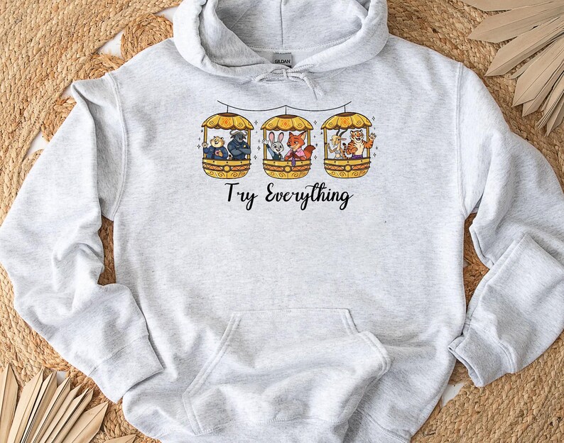 Pode incluir: Moletom cinza claro com bolso canguru e capuz com cord&atilde;o. A frente apresenta um gr&aacute;fico de tr&ecirc;s g&ocirc;ndolas, cada uma com um animal, e as palavras "Try Everything" em escrita preta.
