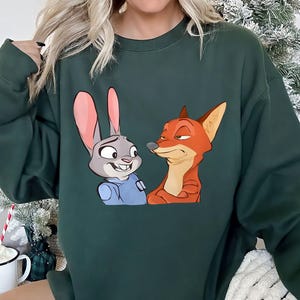 May include: Dark green sweatshirt featuring a cartoon image of Judy Hopps and Nick Wilde from Zootopia. Judy is a gray rabbit wearing a blue uniform, and Nick is an orange fox. The sweatshirt is a solid color.