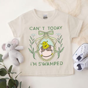 Puede incluir: Una camiseta beige con una ilustraci&oacute;n verde, amarilla y blanca de Shrek en un marco. El texto "Can't today I'm swamped" est&aacute; impreso en la camiseta.