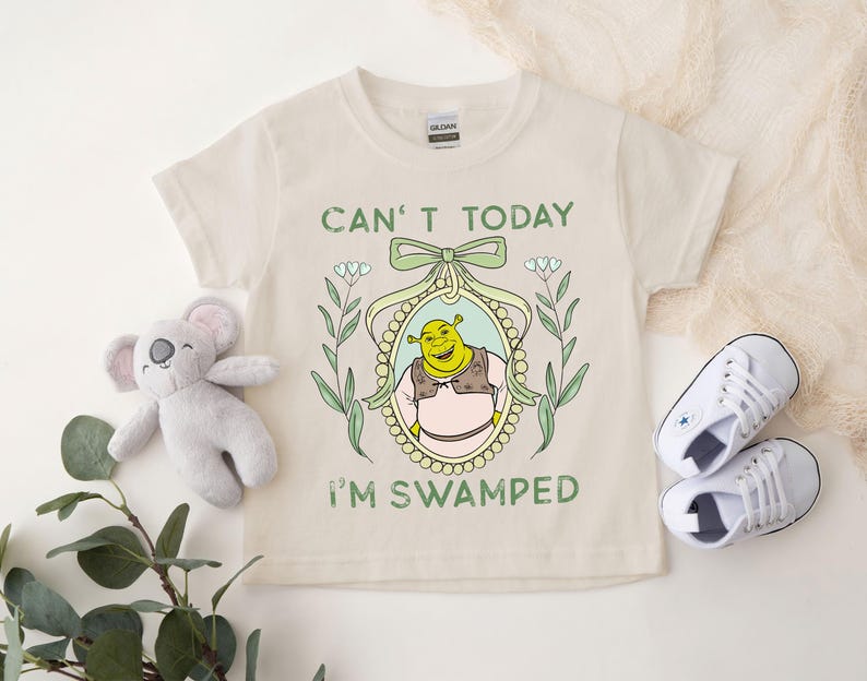 Puede incluir: Una camiseta beige con una ilustraci&oacute;n verde, amarilla y blanca de Shrek en un marco. El texto "Can't today I'm swamped" est&aacute; impreso en la camiseta.