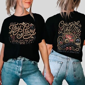 May include: A black t-shirt featuring a graphic design on the front and back. The front displays the text "It's a Toy Story, Baby, just say yet" in rope-style lettering. The back features the text "Cowgirl Like Me" and "Takes One to Know One".