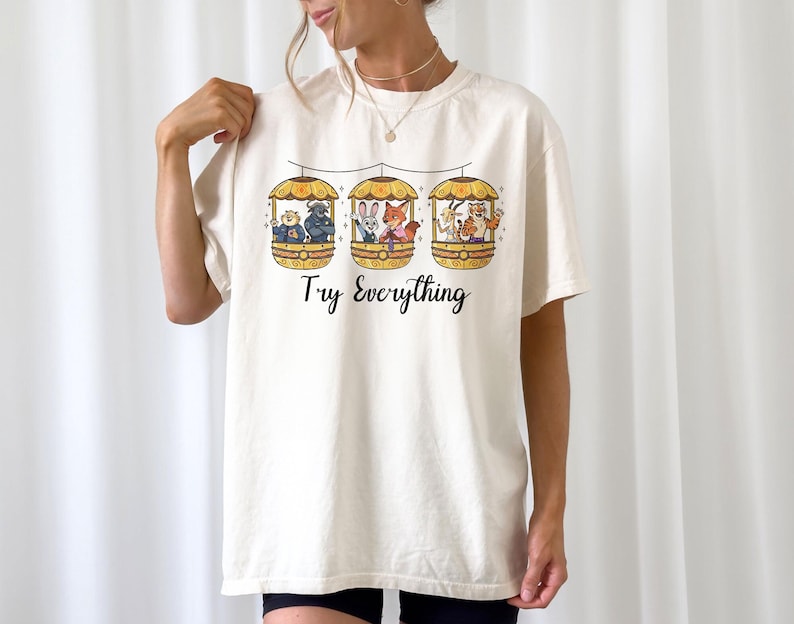 Pode incluir: Camiseta creme com um gr&aacute;fico extravagante de tr&ecirc;s g&ocirc;ndolas, cada uma com animais de desenho animado. As g&ocirc;ndolas est&atilde;o suspensas por uma linha, e o texto "Try Everything" est&aacute; impresso abaixo.