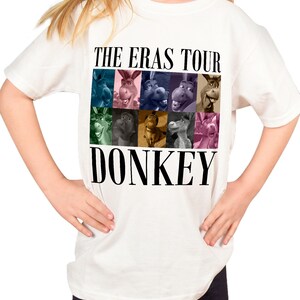 Pode incluir: Camiseta branca com um gr&aacute;fico preto de Donkey de Shrek. O gr&aacute;fico apresenta uma colagem do rosto de Donkey de diferentes filmes da franquia Shrek. Os textos "The Eras Tour" e "Donkey" est&atilde;o impressos em preto.