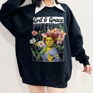 May include: Black sweatshirt featuring a graphic print of a green ogre character wearing a crown of flowers. The text "Grit & Grace" is printed above the character.