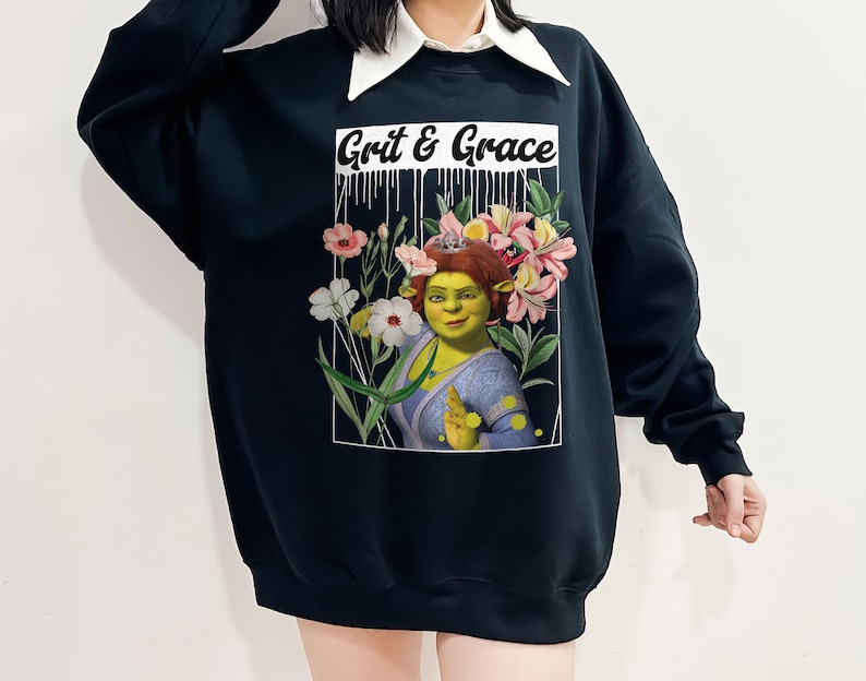May include: Black sweatshirt featuring a graphic print of a green ogre character wearing a crown of flowers. The text "Grit & Grace" is printed above the character.