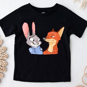 May include: Black short-sleeved t-shirt featuring a cartoon illustration of a rabbit and a fox. The rabbit has gray fur, pink ears, and is wearing a blue shirt. The fox is orange and red. The characters are from the animated movie Zootopia.