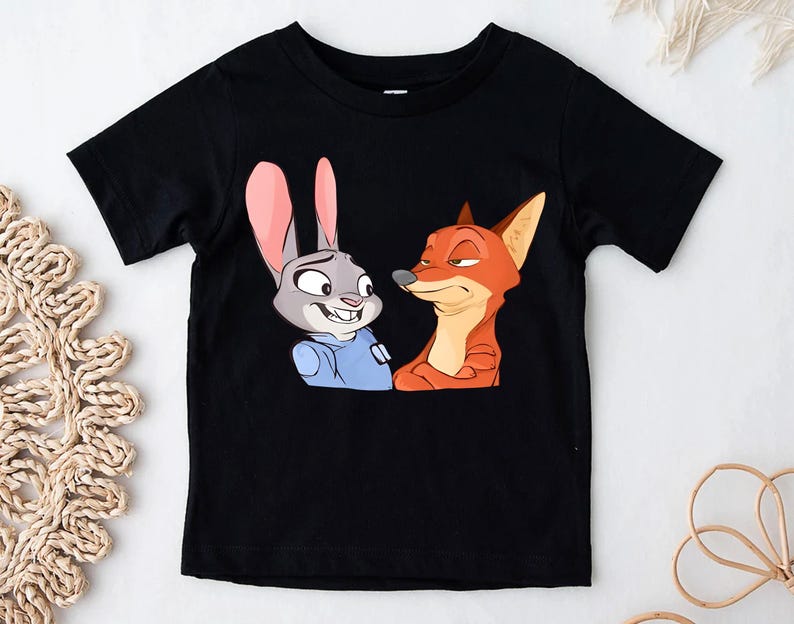 May include: Black short-sleeved t-shirt featuring a cartoon illustration of a rabbit and a fox. The rabbit has gray fur, pink ears, and is wearing a blue shirt. The fox is orange and red. The characters are from the animated movie Zootopia.