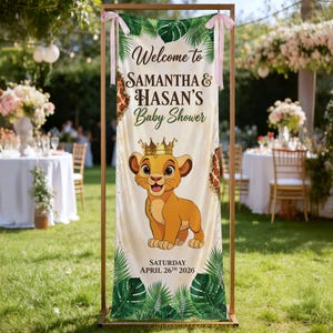 May include: A vertical banner featuring a cartoon lion cub wearing a gold crown, surrounded by tropical leaves and giraffe print accents. The text reads: Welcome to SAMANTHA & HASAN'S Baby Shower, SATURDAY APRIL 26TH 2026. The banner is held by pink ribbons.